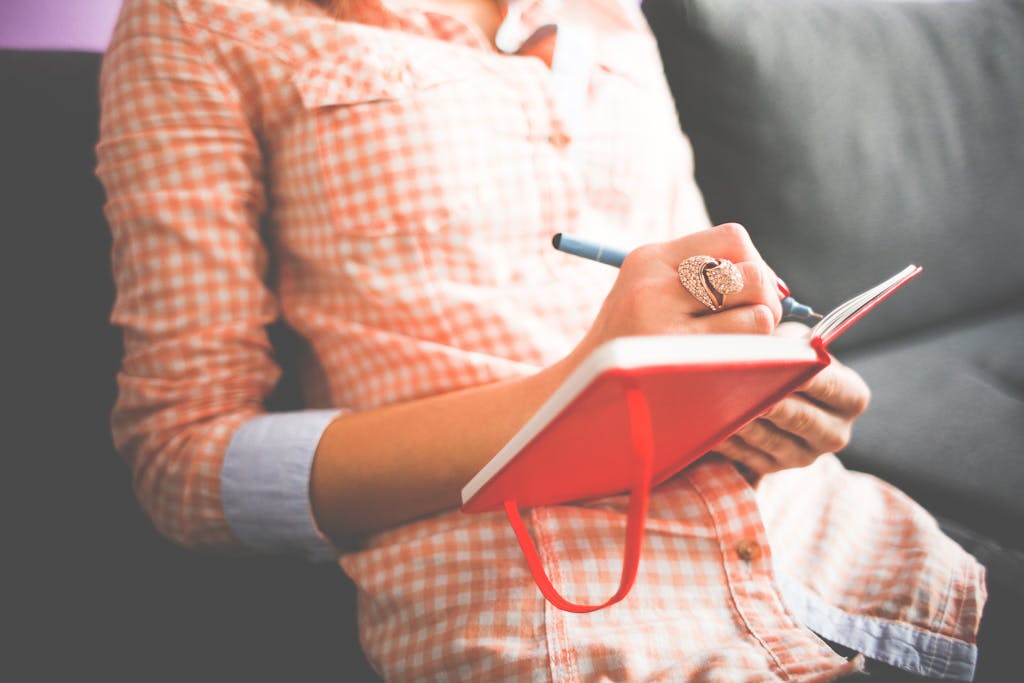 Close-up of a woman writing in a red journal on a sofa, wearing a stylish ring.